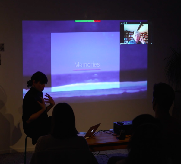 A photograph of Jesssica Scott performing seated with a projected presentation in front of a crowd.