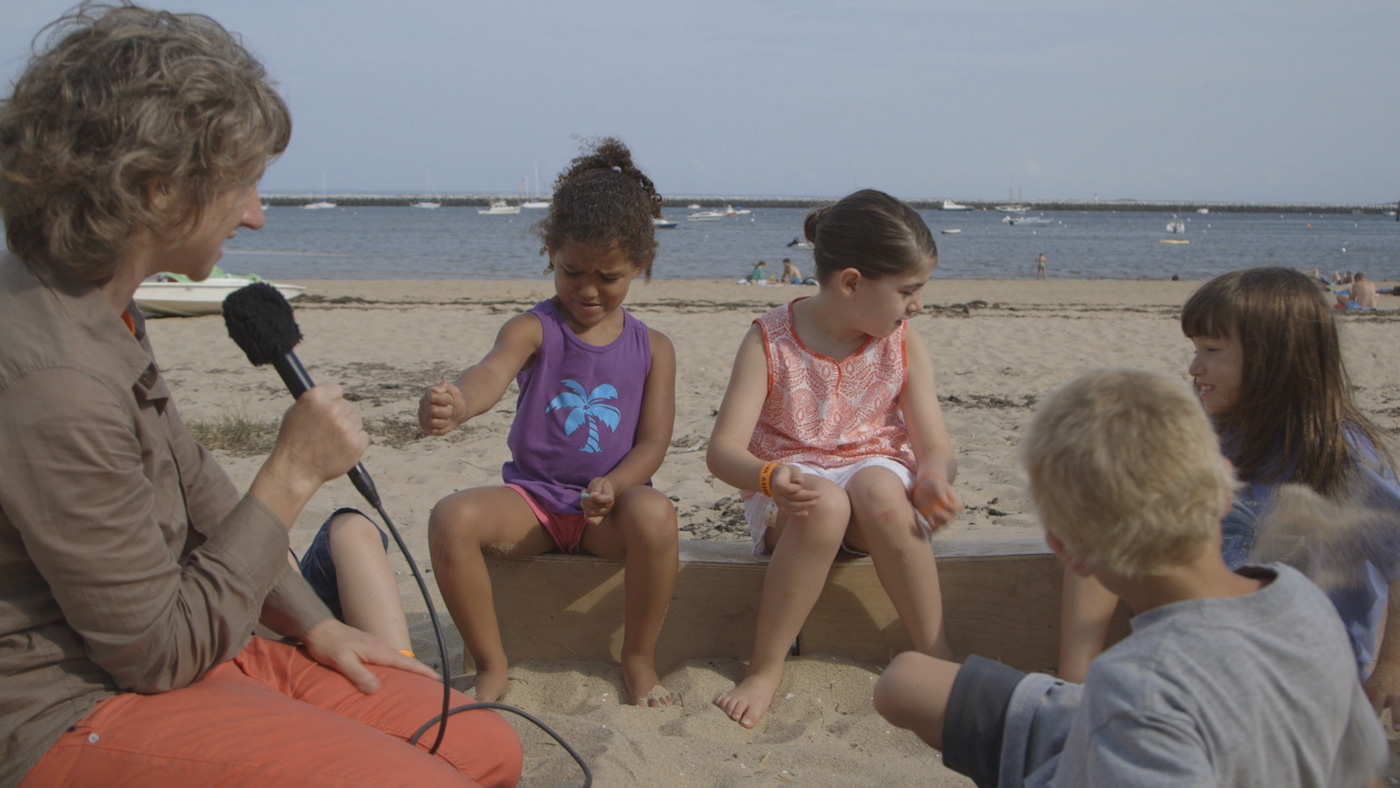 video still of a group of children being interviewed by an adult woman at the beach